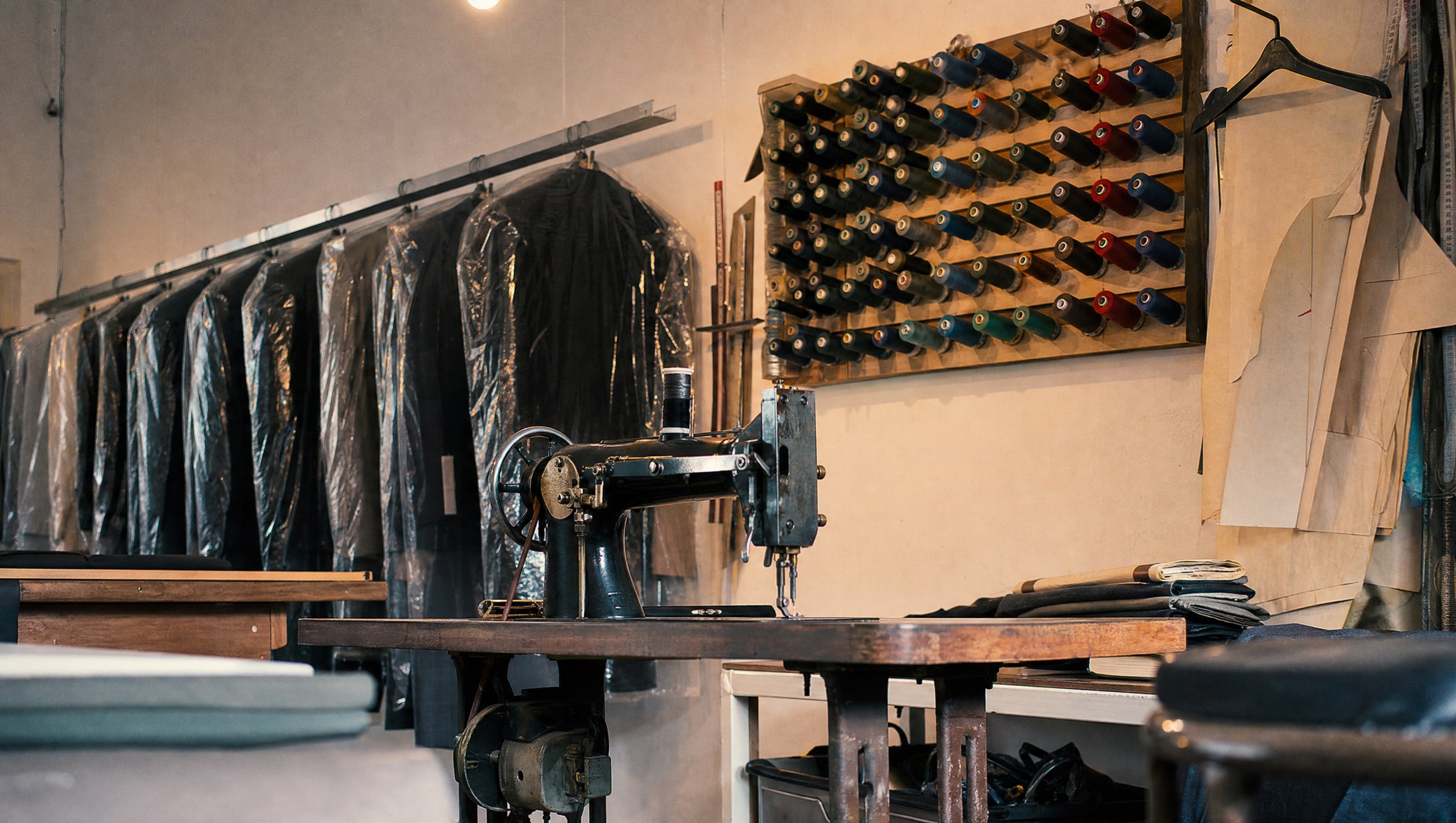 Inside the workshop. A row of finished jackets in plastic on the rail, a Singer industrial sewing machine on the table, a wooden threadboard on the wall with rows of coloured spools.